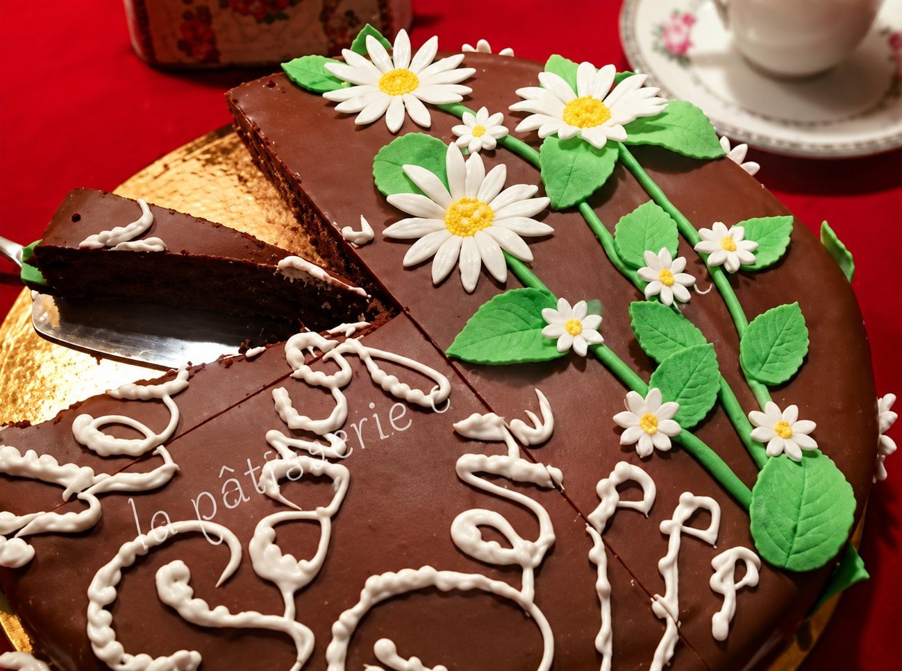 Gâteau au chocolat orné de marguerites et de feuilles en pâte à sucre, avec une inscription en glaçage blanc.